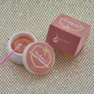 Small round container with lid placed to the side revealing the peach colored cream inside. Small micro brush near product. Peach colored box package in background. Items against golden texture background. 
