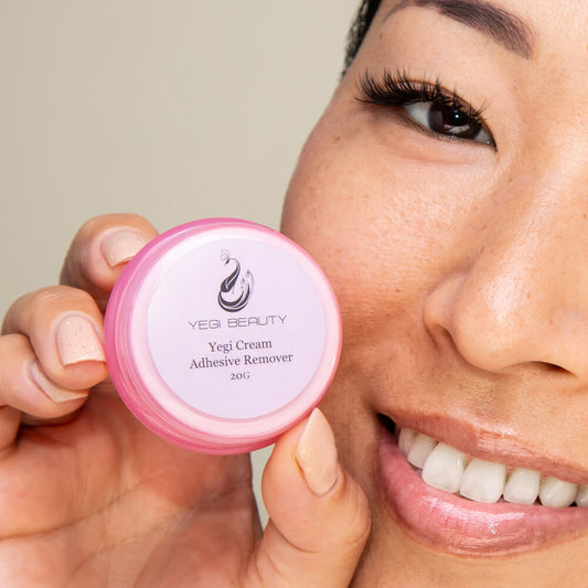 20g tiny pink round container with cap off and placed to the side revealing orange cream inside. Cap label reads "Yegi Cream Adhesive Remover." Product held by smiling woman very close to face to show eyelash extensions 