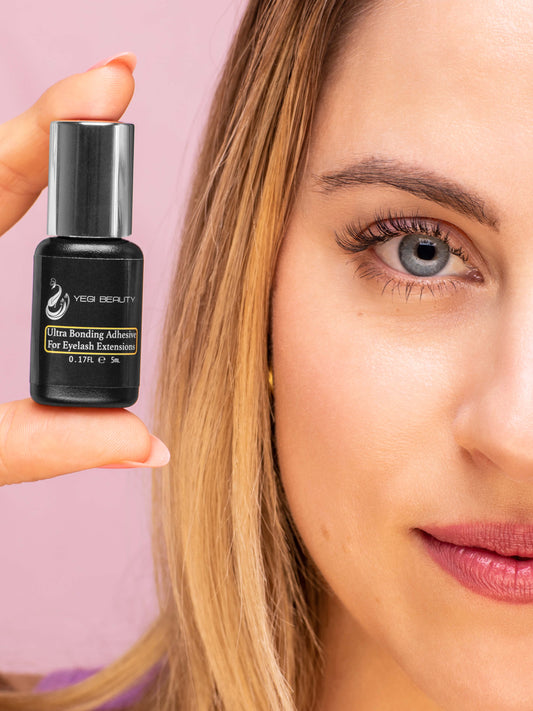 5ml black bottle with silver cap against white background. Label with yellow rectangle reads "Ultra Bonding Adhesive For Eyelash Extensions." Yellow indicates this is banana scented. Held between thumb and finger by woman against pink background. Woman sporting classic eyelash extensions