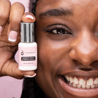 Closeup image of woman smiling holding a small bottle next to her cheek. Bottle reads "Mega Hold Adhesive for eyelash extensions" against pink background.