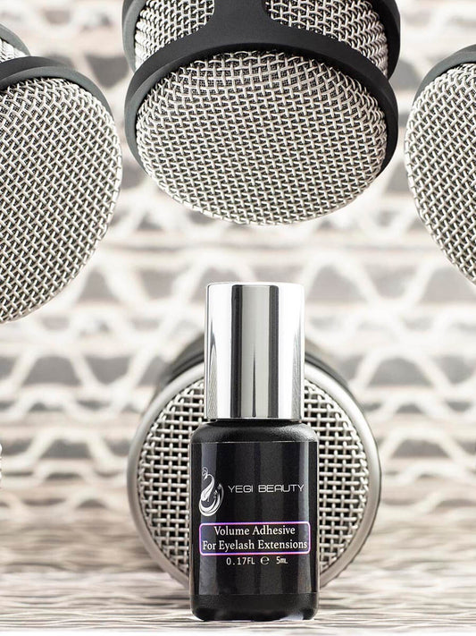 5ml black bottle with silver cap. Label reads "Yegi Beauty Volume Adhesive For Eyelash Extensions" with purple rectangle around words. Against background of microphones to emphasize the idea of volume. 