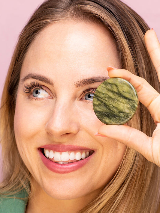 Beautiful woman smiling with classic eyelash extensions holding a green jade stone close to her face. 